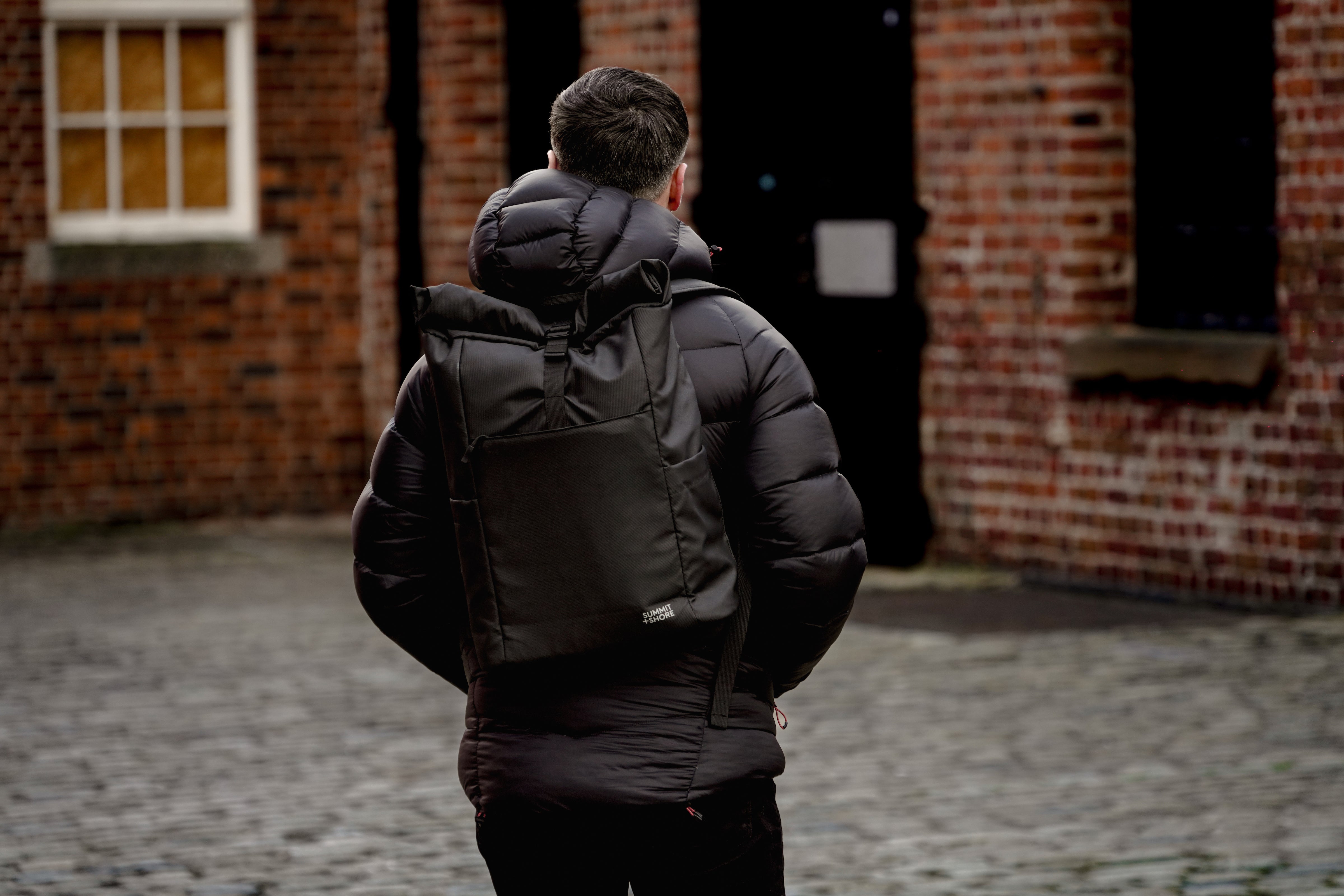 All Black Outfit with Summit and Shore Roll Top Backpack in a cobbled red brick industrial backdrop
